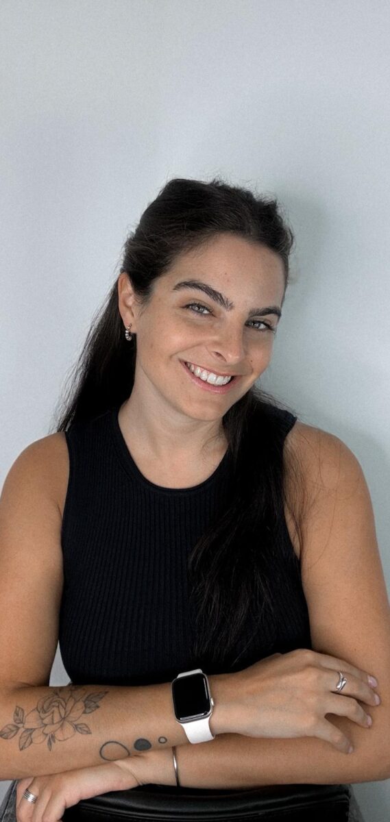 Mujer sonriendo con cabello largo y oscuro, vistiendo una camiseta sin mangas negra, con tatuajes visibles en el brazo. Lleva un reloj inteligente en la muñeca y está posando con los brazos cruzados frente a un fondo blanco.