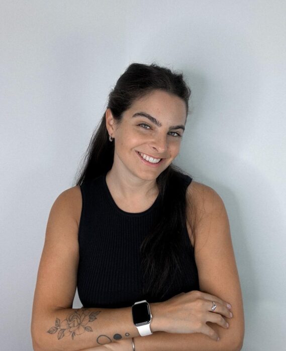 Mujer sonriendo con cabello largo y oscuro, vistiendo una camiseta sin mangas negra, con tatuajes visibles en el brazo. Lleva un reloj inteligente en la muñeca y está posando con los brazos cruzados frente a un fondo blanco.