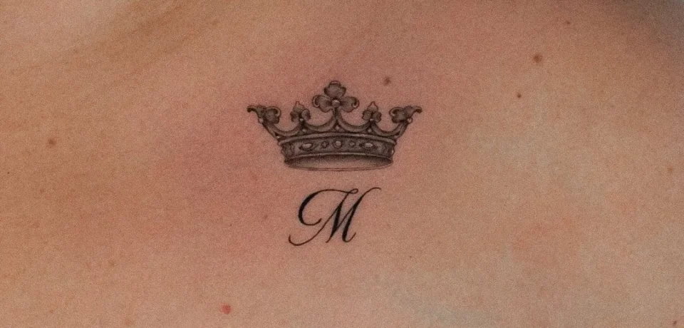 Tatuaje de una corona con la letra "M" en el hombro de una persona, con un diseño detallado y elegante.