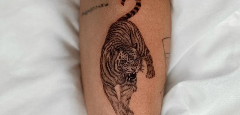 Tatuaje de un tigre corriendo en el antebrazo, con detalles realistas y expresión feroz, sobre un fondo de sábanas blancas.